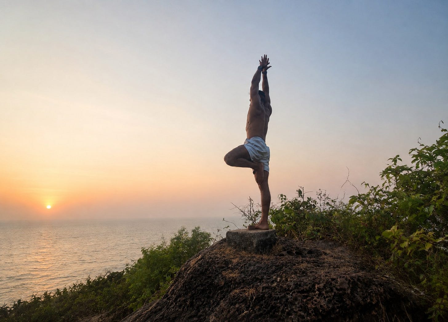 Avi in tree pose on a rock at sunset, arms raised, sea in the background Avi in tree pose on a rock at sunset, arms raised, sea in the background