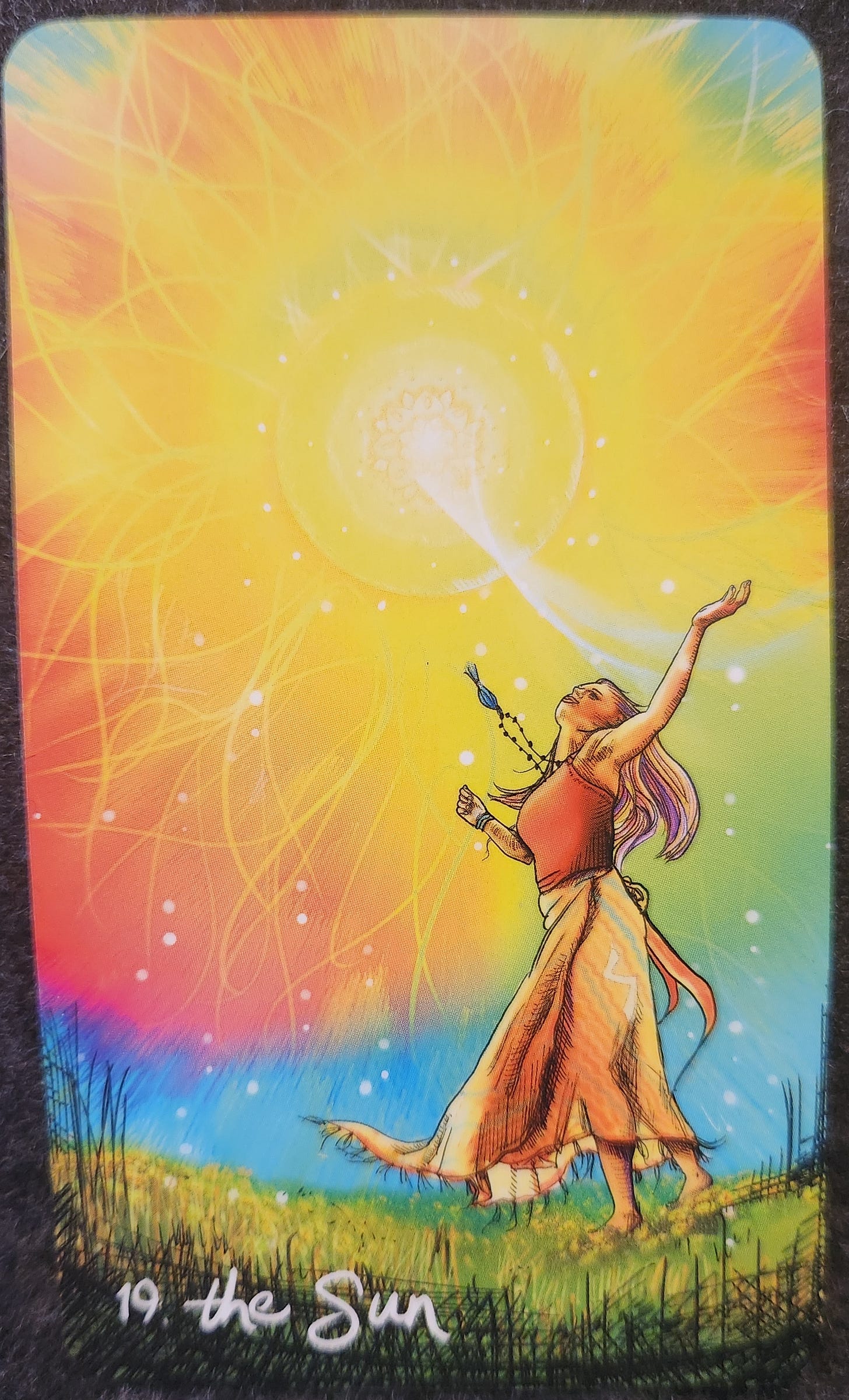 A tarot card labelled 19. The Sun. A woman is standing in a field dressed in a red top and long orange skirt. Her arms are opened wide, her chest and face tilted towards the sky. The sun in the sky is burning brightly, surrounded by rays of light that appear as flower petals. A beam of sunlight is shooting down from the sun to the woman’s chest. 