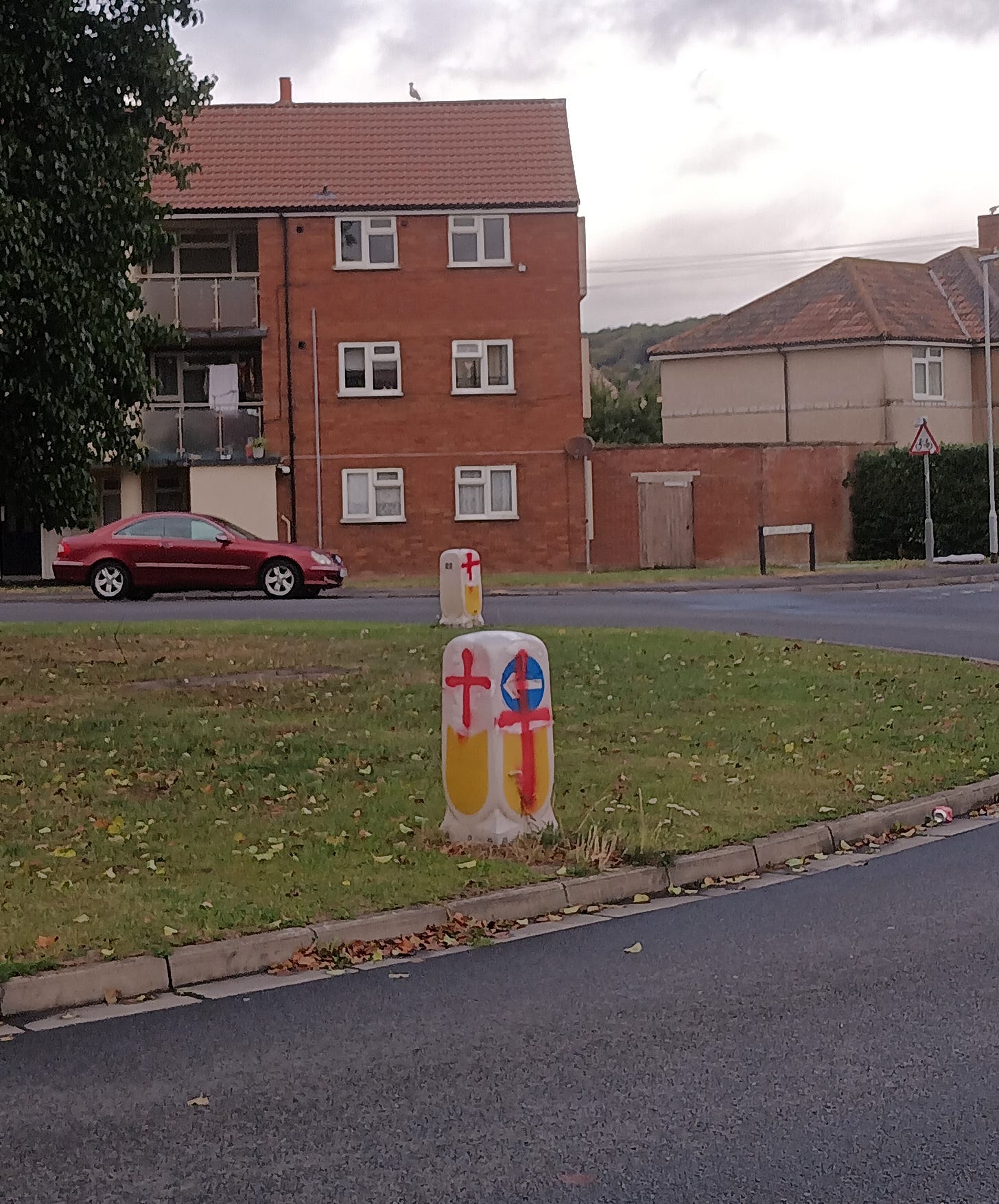 A traffic bollard, which is mostly yellow and blue with some patches of white. Someone has still tried to paint a red cross over it, which hasn't really worked out, what with all the yellow and blue sections getting in the way.