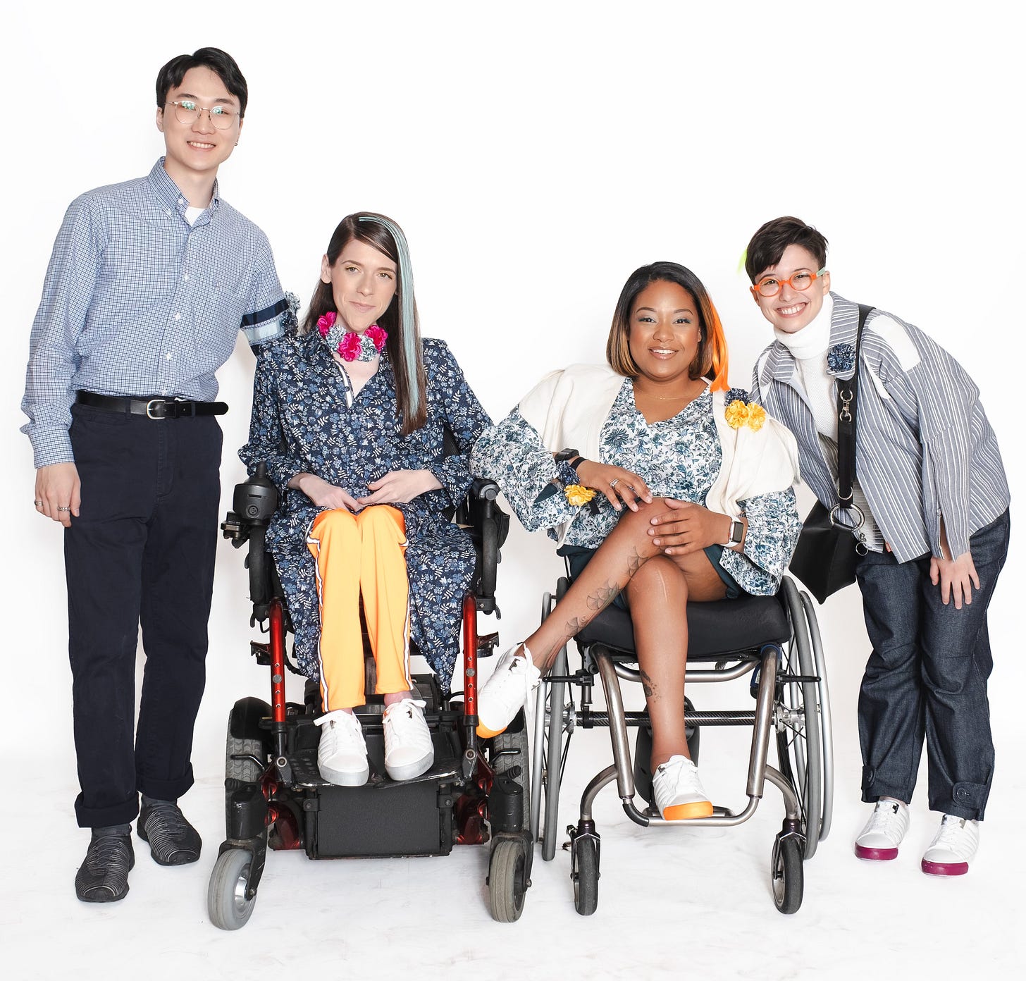 A warm group portrait of four stylish models and designers against a white background. Two women are seated in wheelchairs at the center of the group, smiling warmly. They are flanked by a standing man and another person who is leaning in, creating a sense of camaraderie and teamwork.