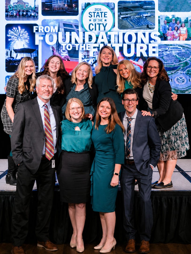 The City of Dublin, Ohio communications team stands together on a stage, smiling at the camera. The group includes men and women dressed in business attire, arranged in two rows. Behind them, a large screen displays “2026 State of the City: From Foundations to the Future,” along with images of city projects and community scenes.