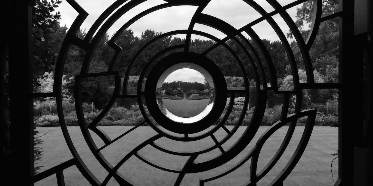 GPT-4o: A black and white photograph features an ornate metal gate with an intricate circular design composed of interconnected arcs and angular shapes. At the center of the gate is a circular lens that provides a fish-eye view of a garden beyond. Through the lens, the viewer sees a distorted, wide-angle perspective of a manicured lawn, flowerbeds, and dense trees in the background. The gate’s geometric framework surrounds the lens, drawing attention to the central focal point while offering glimpses of the garden through its open spaces. The composition creates a striking balance between the architectural structure of the gate and the organic beauty of the garden beyond.