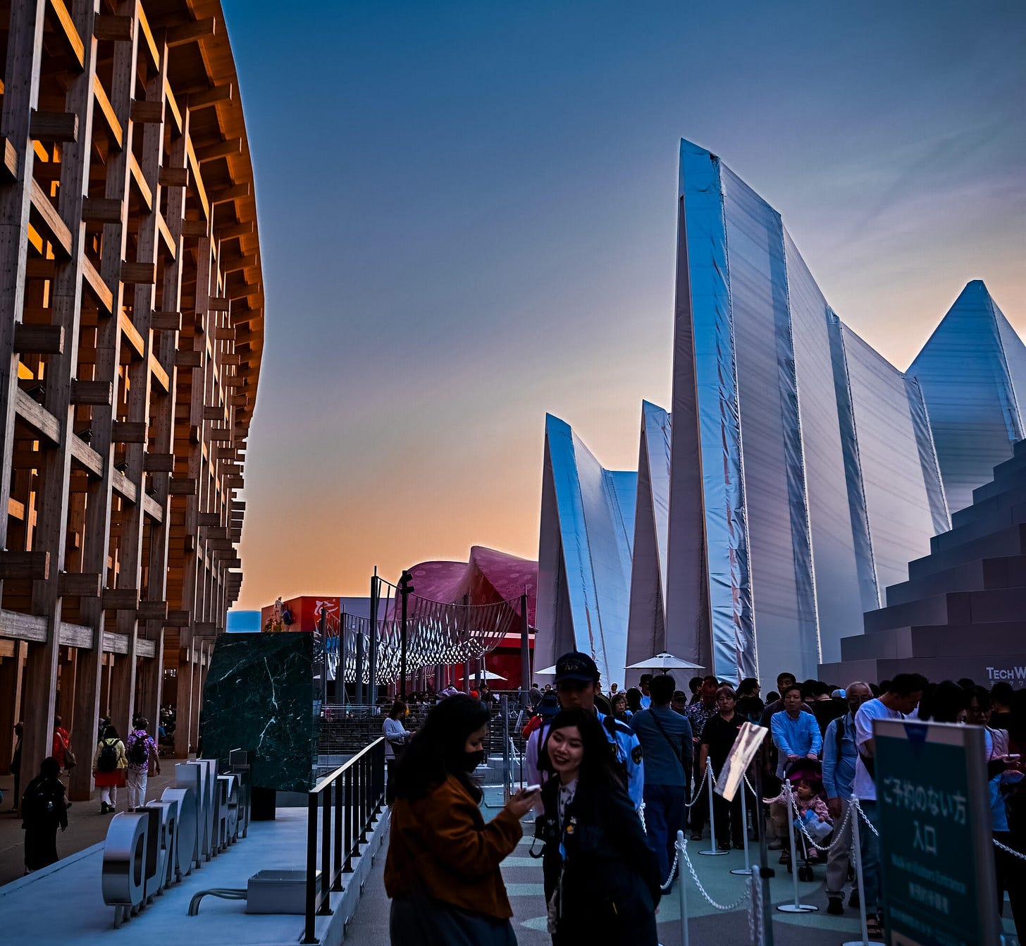 Crowds line up between a multi-story curved wooden structure and metallic pavilions at the World Expo at sunset
