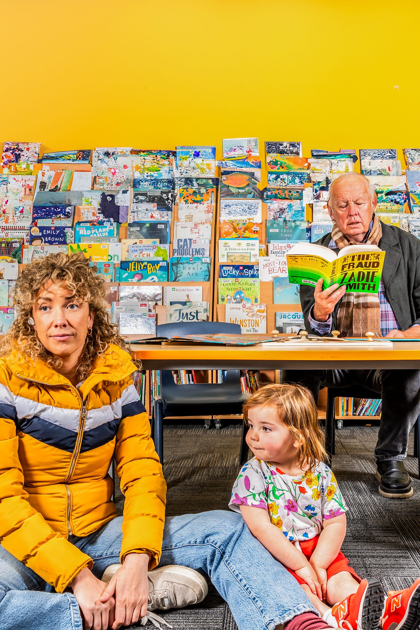 A woman in a yellow puffer jacket and jeans sits on the floor next to a small child with blonde hair wearing a patterned t-shirt. The woman looks toward the camera with a weary expression, while the child looks to the side. Behind them, a man in a blazer and scarf sits at a table reading a bright green book. The background wall is bright yellow and covered halfway up with a colorful, dense display of children’s books.