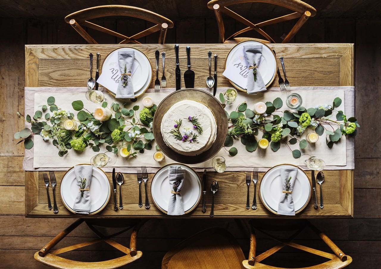 Overhead view of an intimate table setting for five, with white plates, candles, greenery, and a small cake at the center. Overhead view of an intimate table setting for five, with white plates, candles, greenery, and a small cake at the center.