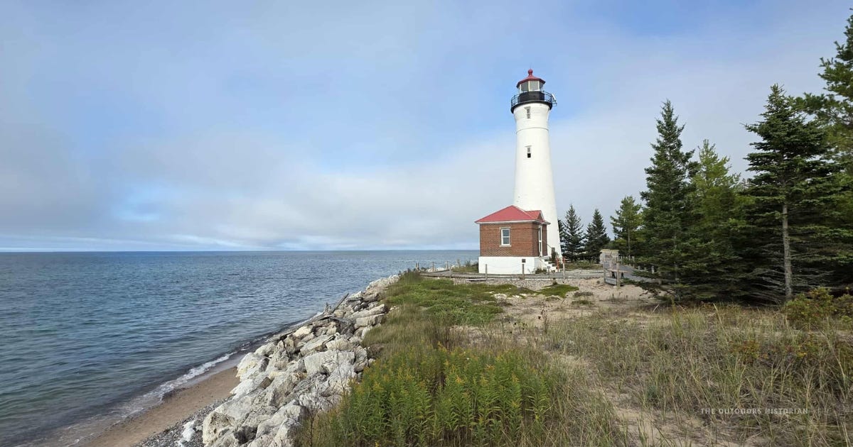 Crisp Point Lighthouse, a white tower with a red roof, standing along a rocky Lake Superior shoreline with dune grass in the foreground.