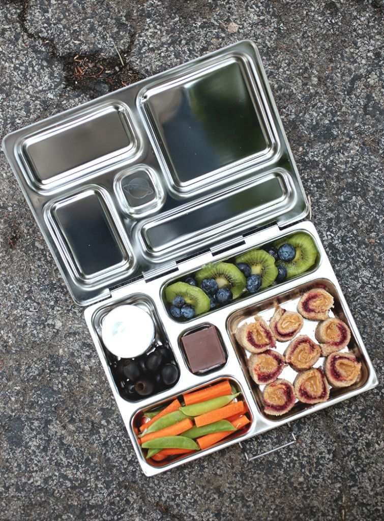 An open stainless PlanetBox lunch box packed with food, showing separate compartments and a small sealed container inside. An open stainless PlanetBox lunch box packed with food, showing separate compartments and a small sealed container inside.