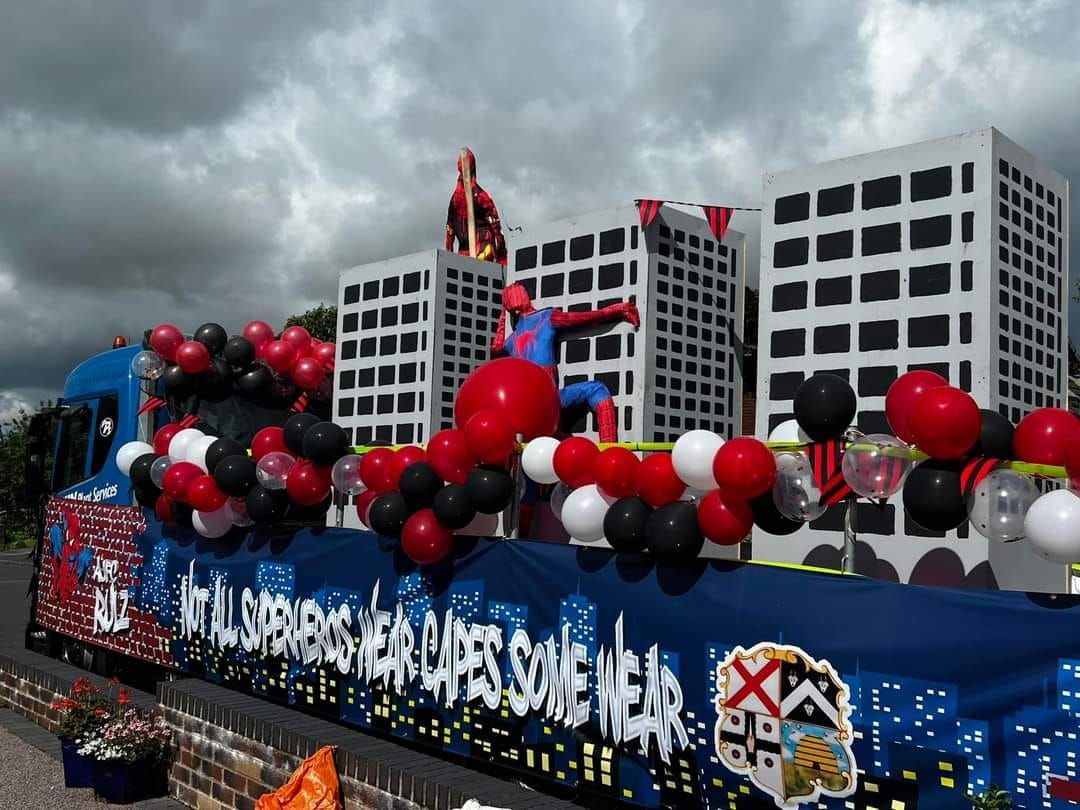 A carnival float with a blue and red Spiderman superhero climbing grey sky scrapers on the back.
