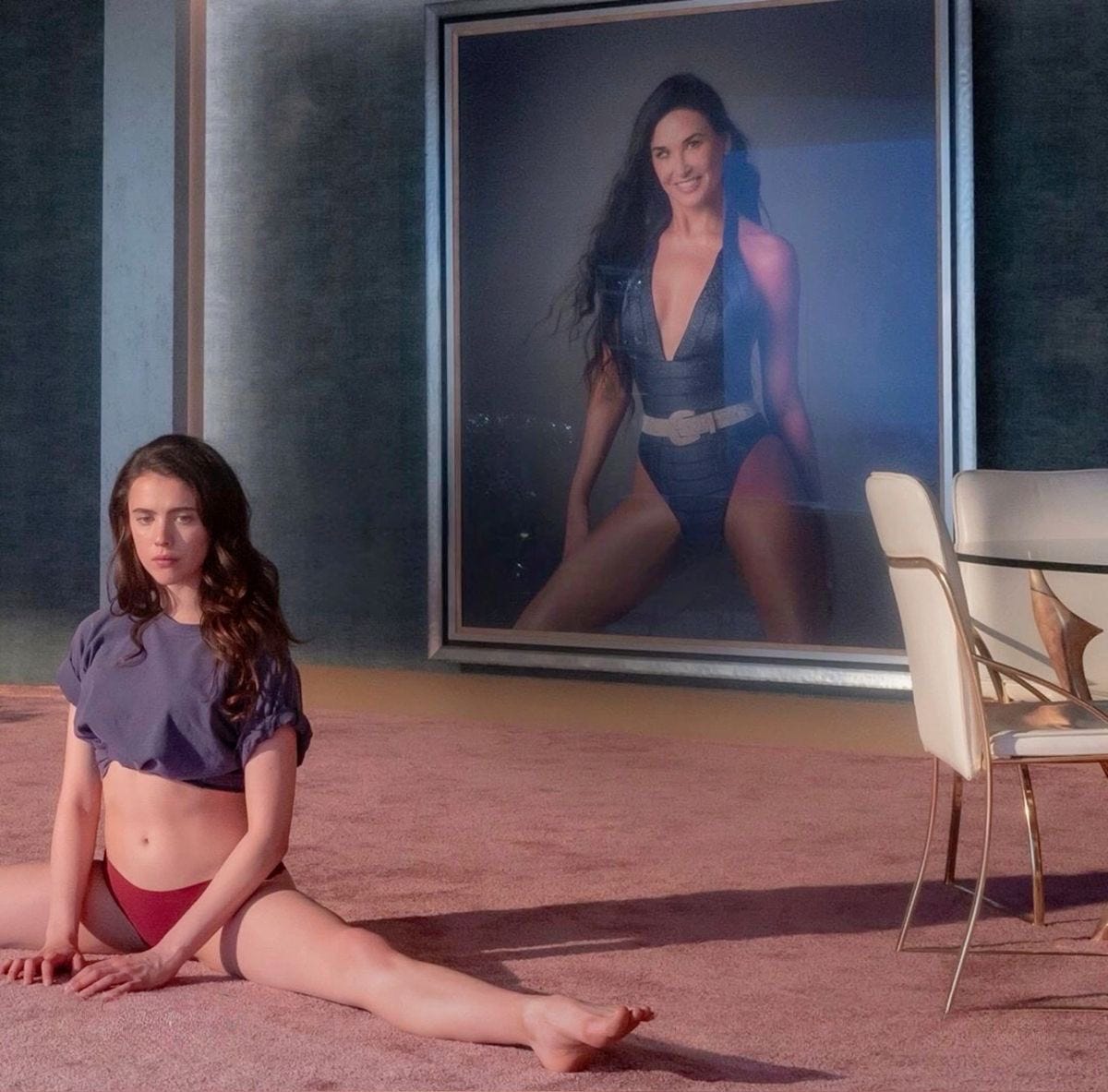 Cena do filme A substância. No chão, uma mulher jovem de calcinha e camiseta está sentada com as pernas esticadas. No fundo da foto, é possível ver um retrato pendurado na parede, de uma mulher de 50 e poucos anos, com uma roupa também sensual.