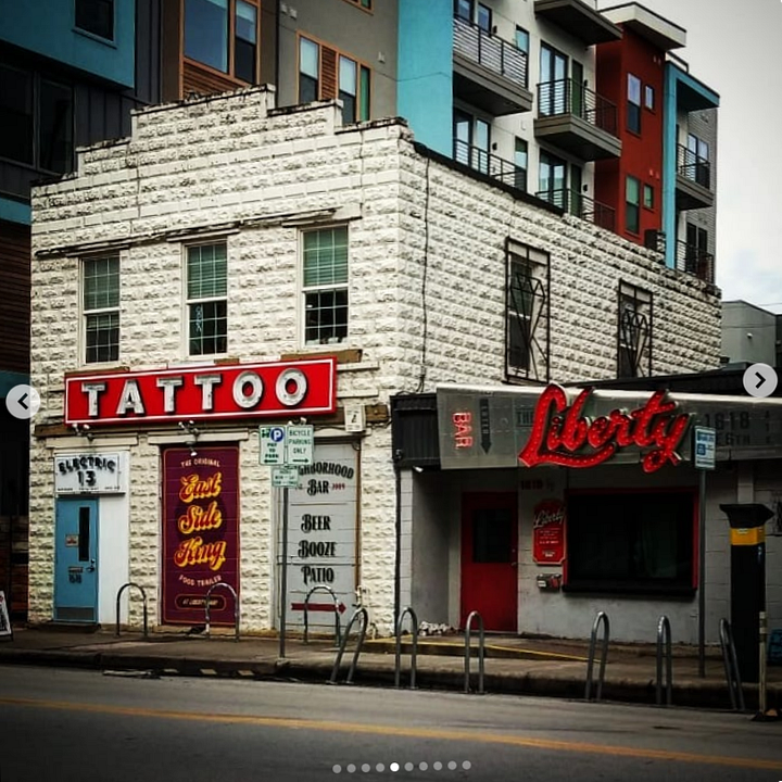 1. a white building that looks like a house, with "Church of God of the Firstborn" written on it 2. the back of a street sign, covered in band stickers and I.C.E. out of Texas sticker. 3. the toes of my boots next to a manhole cover with rings of hexagons stamped into it. The Liberty Bar, and a tattoo parlour in 100+ year old buildings surrounded by newer, taller buildings