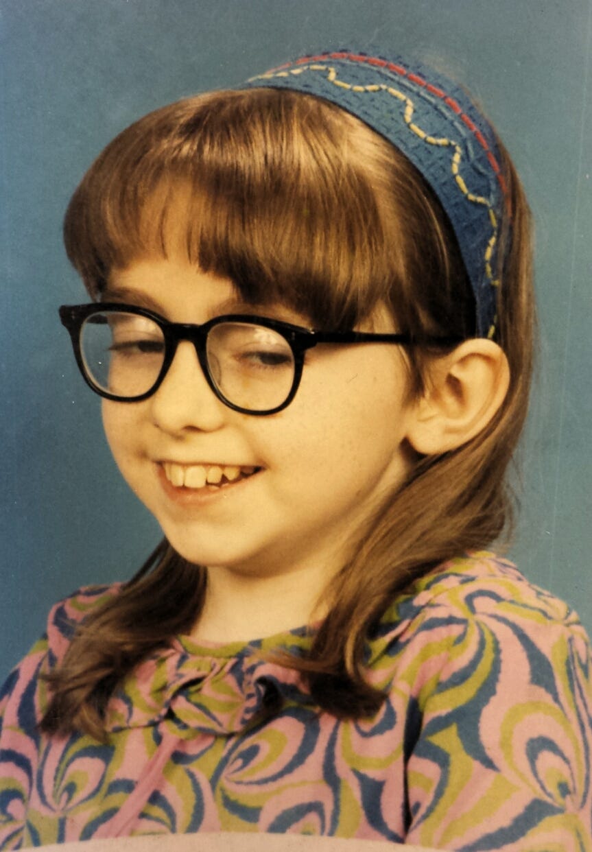 A school photo of Penny from 1971 when she was 10 years old. Penny has nut-brown hair, shoulder length kept behind her ears. She wears a homemade embroidered blue headband and has a toothy grin. Her glasses are old-school NHS black frames. She is smiling.