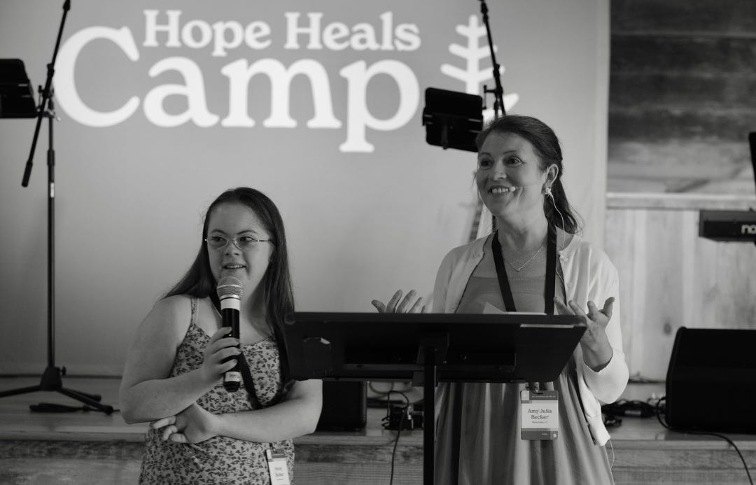 A black and white photo by Bryan Johnson of Penny and me standing near a wooden stage under a sign that reads "Hope Heals Camp." 