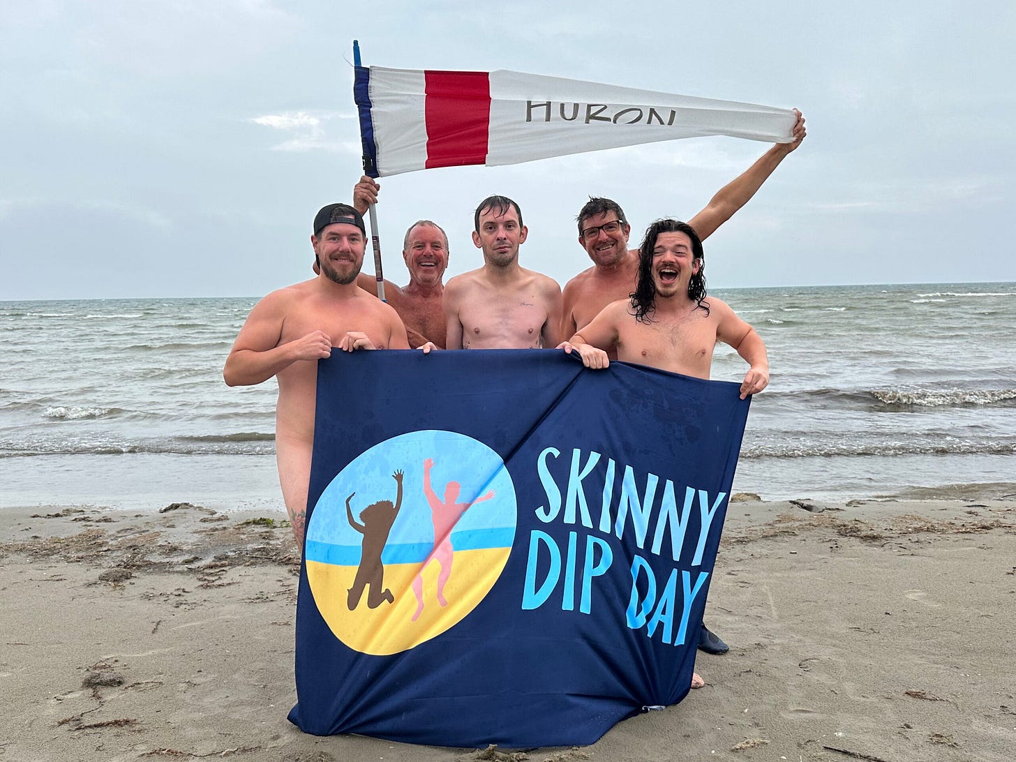Five nude people pose on a beach holding a Skinny Dip Day banner, with one member behind them raising a white flag labeled "HURON." Waves from Lake Huron roll in behind them under a cloudy sky. Five nude people pose on a beach holding a Skinny Dip Day banner, with one member behind them raising a white flag labeled "HURON." Waves from Lake Huron roll in behind them under a cloudy sky.