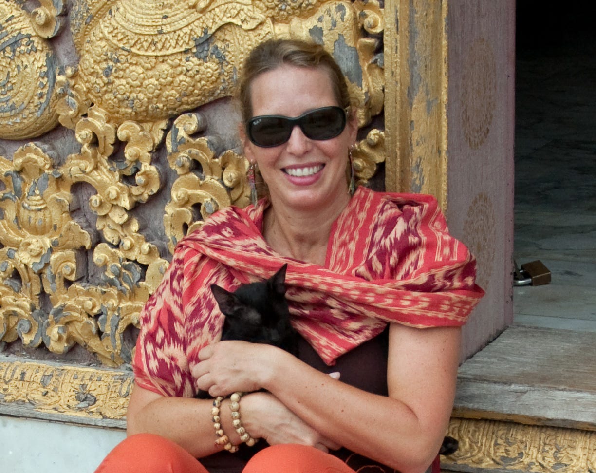 White woman wearing dark sunglasses and a orange and red shawl sitting outside gold ornate template. She is holding a small black kitten in her arms.