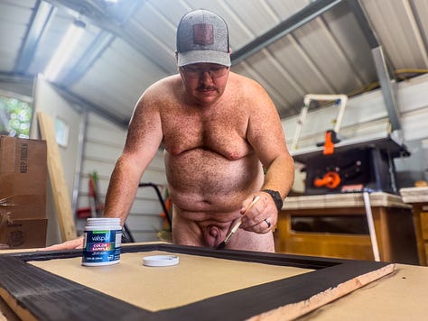 Author Dustin Cox is seen working completely nude in his wood shop. Cutting wood on a saw in the first image. Painting a wood frame black in the second image. Posing with a wood planter box that he built.
