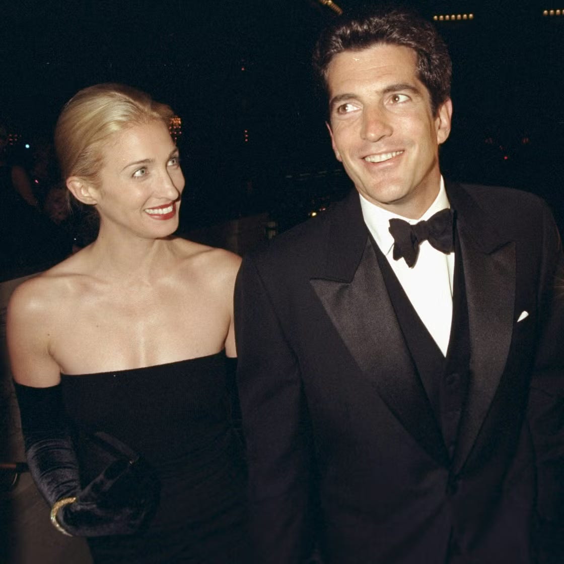 Carolyn Bessette-Kennedy and John F. Kennedy Jr. in black-tie attire; Carolyn wearing a strapless black dress with velvet opera gloves, slicked-back hair, and a signature bold red lip, exemplifying 90s Manhattan evening glamour.