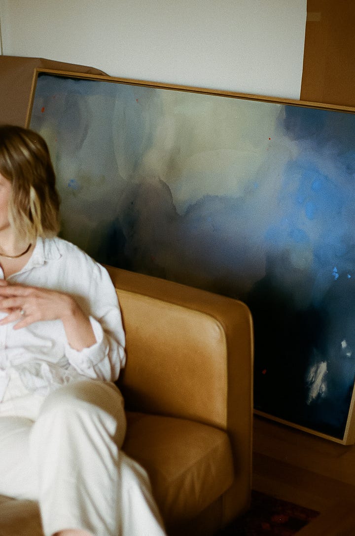 abstract artist Brenna Hodges on a leather couch with her painting in the background; blue acrylic paint dripping on raw canvas