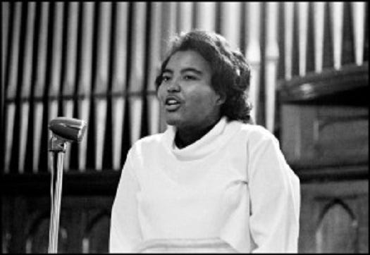 Black and white photo of Prathia Hall in a pulpit with microphone in front of her. She is dressed in all white and preaching