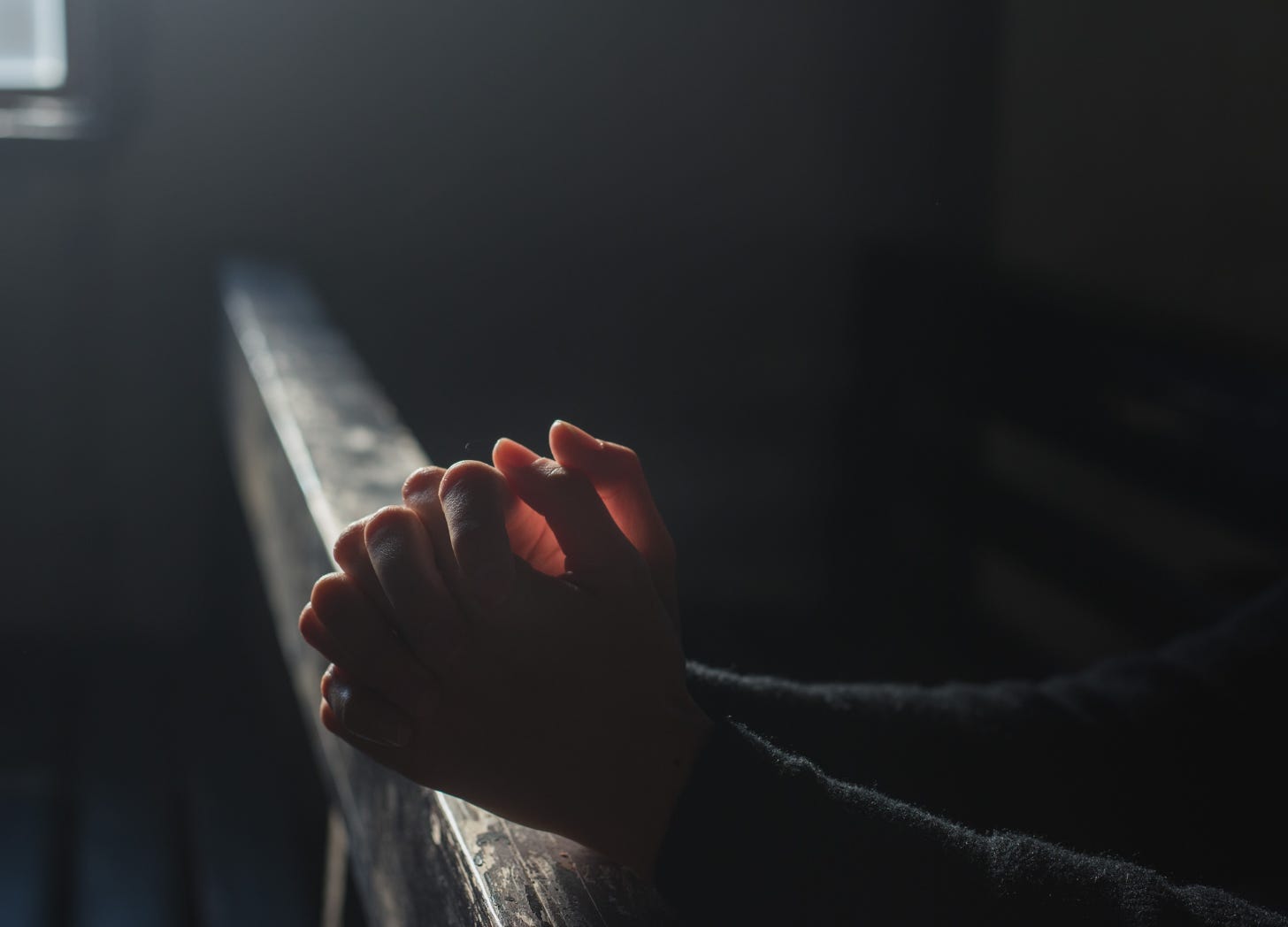 Hands resting in a prayer position on the back of a wooden pew, in a dimly lit church. A soft beam of light shines through a nearby window, illuminating the quiet space.