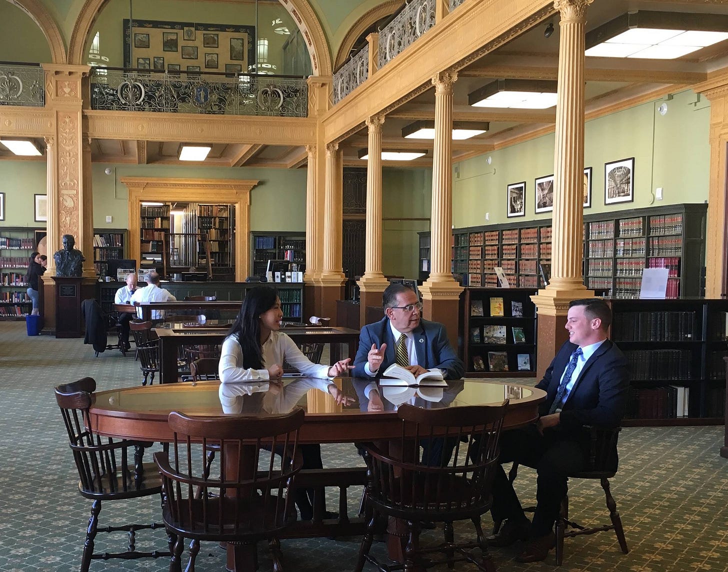 Three people in business attire sit at a round wooden table in a historic library with ornate columns and bookshelves. They appear to be in discussion, with an open book in front of them.