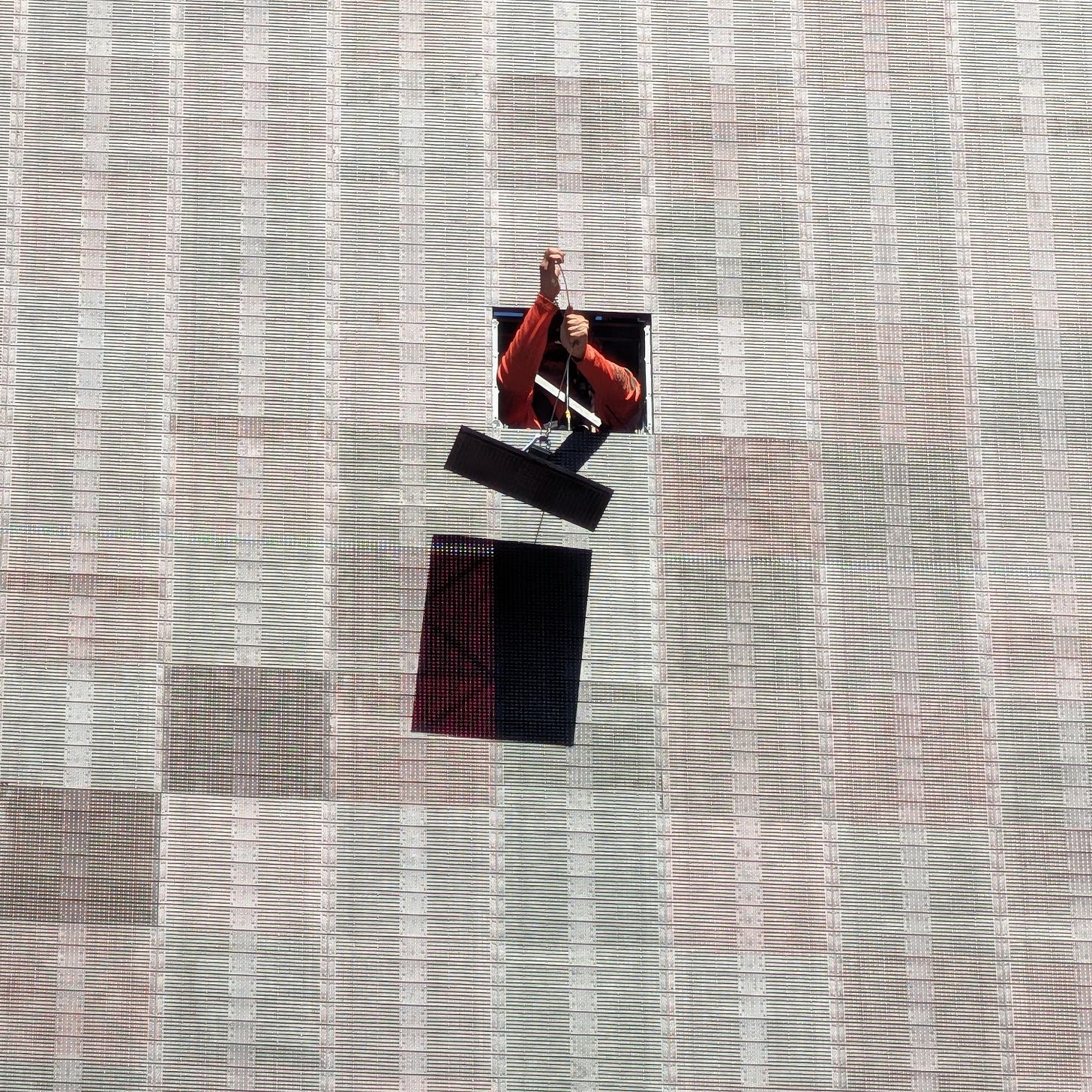 A technician, wearing a bright orange long-sleeved shirt, is positioned behind the LED wall. A rectangular LED panel/module is suspended just below the opening, hanging slightly askew, suggesting it is either being replaced or was just detached.