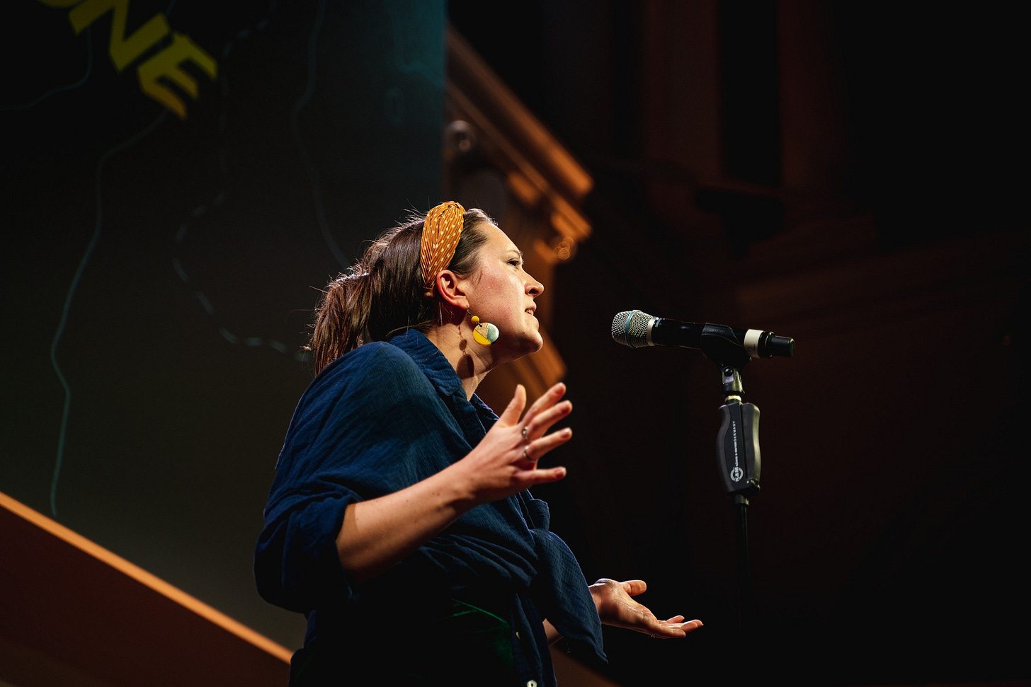 Writer Natalie Jayne Clark is performing on stage at Edinburgh International Book Festival 2023. She is wearing a bright yellow headband and a baggy blue shirt. Her hands are open wide and she has a serious expression on her face as she performs her poetry.