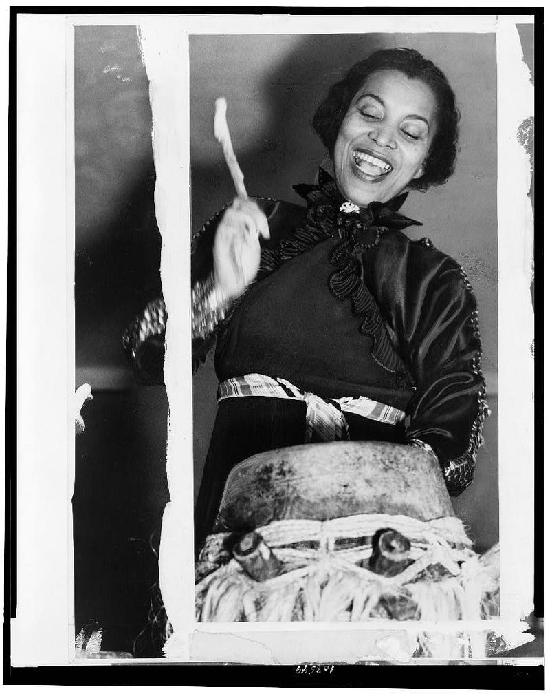 Zora neale hurston wearing a black dress, smiling and beating on an african drum with drumsticks