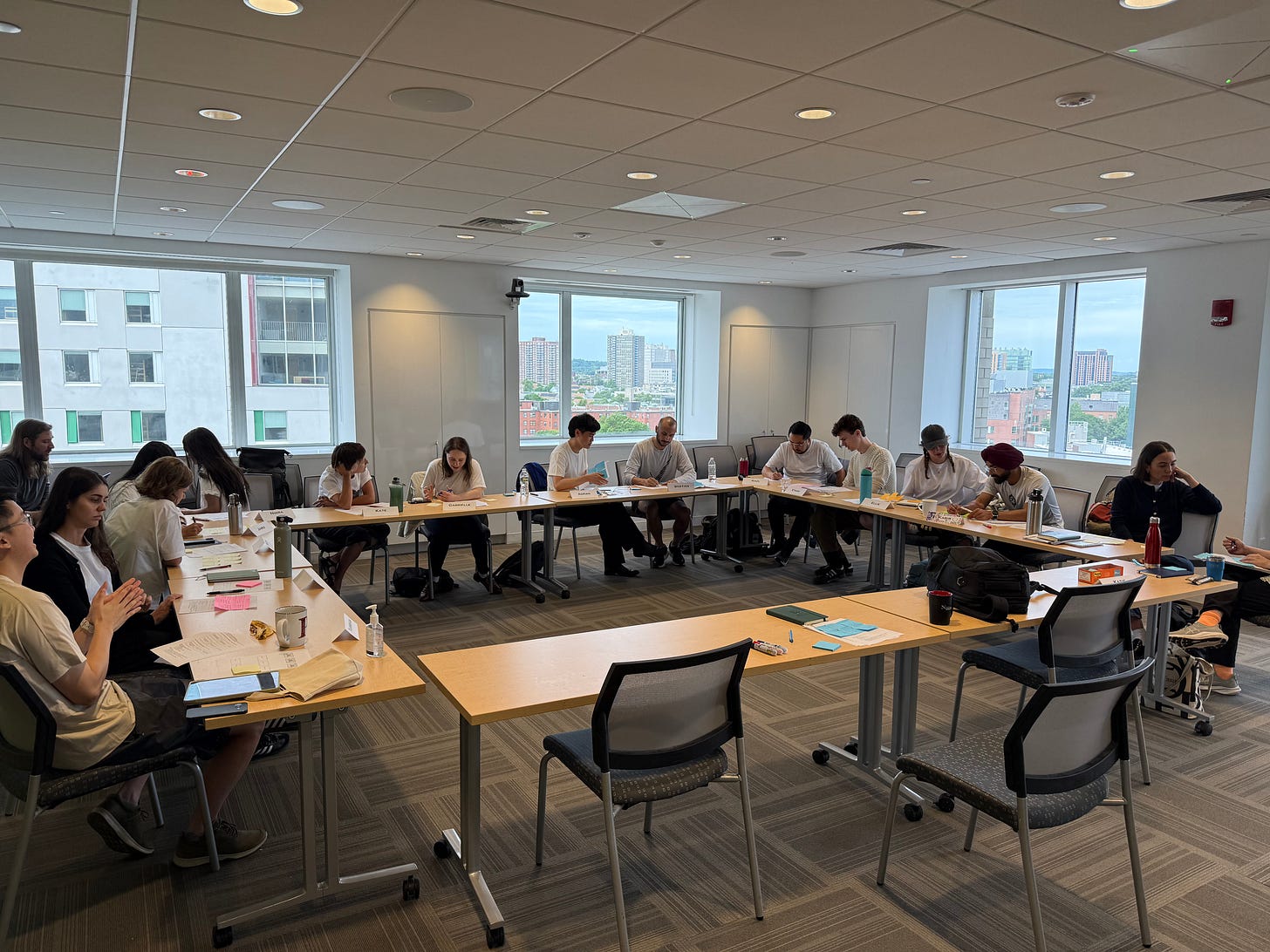 A group of people sit around long tables configured in the shape of a square. They are engaged in conversation, and are reviewing various papers. 