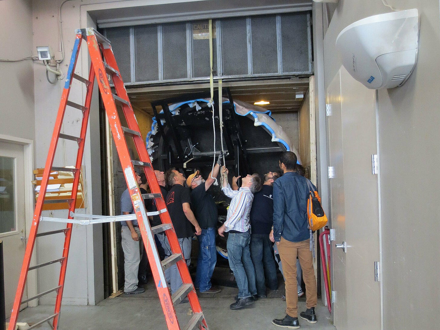 A group of people working together to guide a large object into a freight elevator, with a ladder in front.