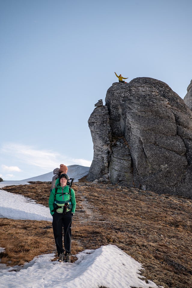 A person in a green jacket stands on tundra below a large rounded granite outcrop, while another person stands atop the rock with arms raised, snow patches around them in warm light.