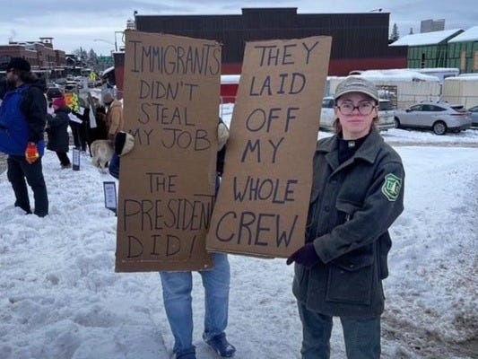 Two Ex-Government employees holding signs that say "Immigrants didn't steal my job, the president did and They laid off my whole crew."