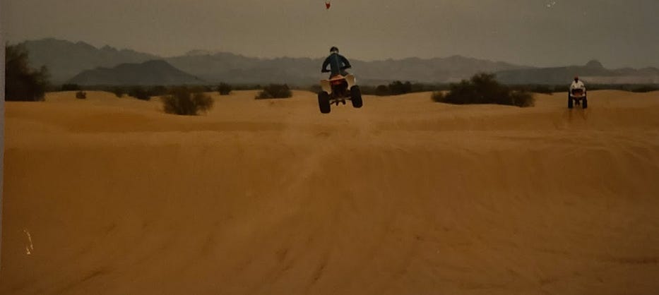 Photo of two riders jumping their ATVs on small sand dunes. One rider is paused and watching the other sail a few feet in the air.