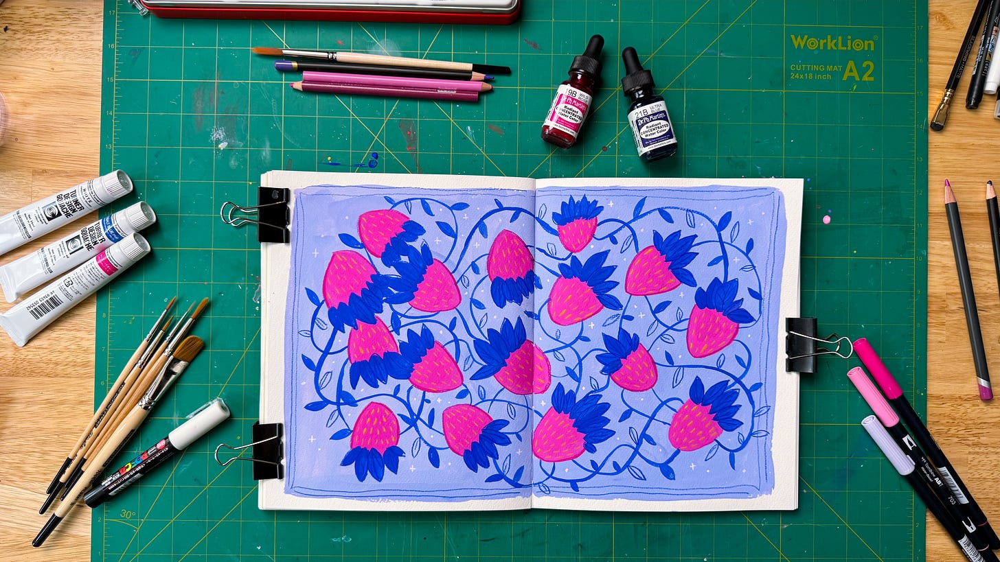 A flatlay of a desk with a sketchbook open to a painting of pink strawberries on a blue background. Various art supplies surround the sketchbook