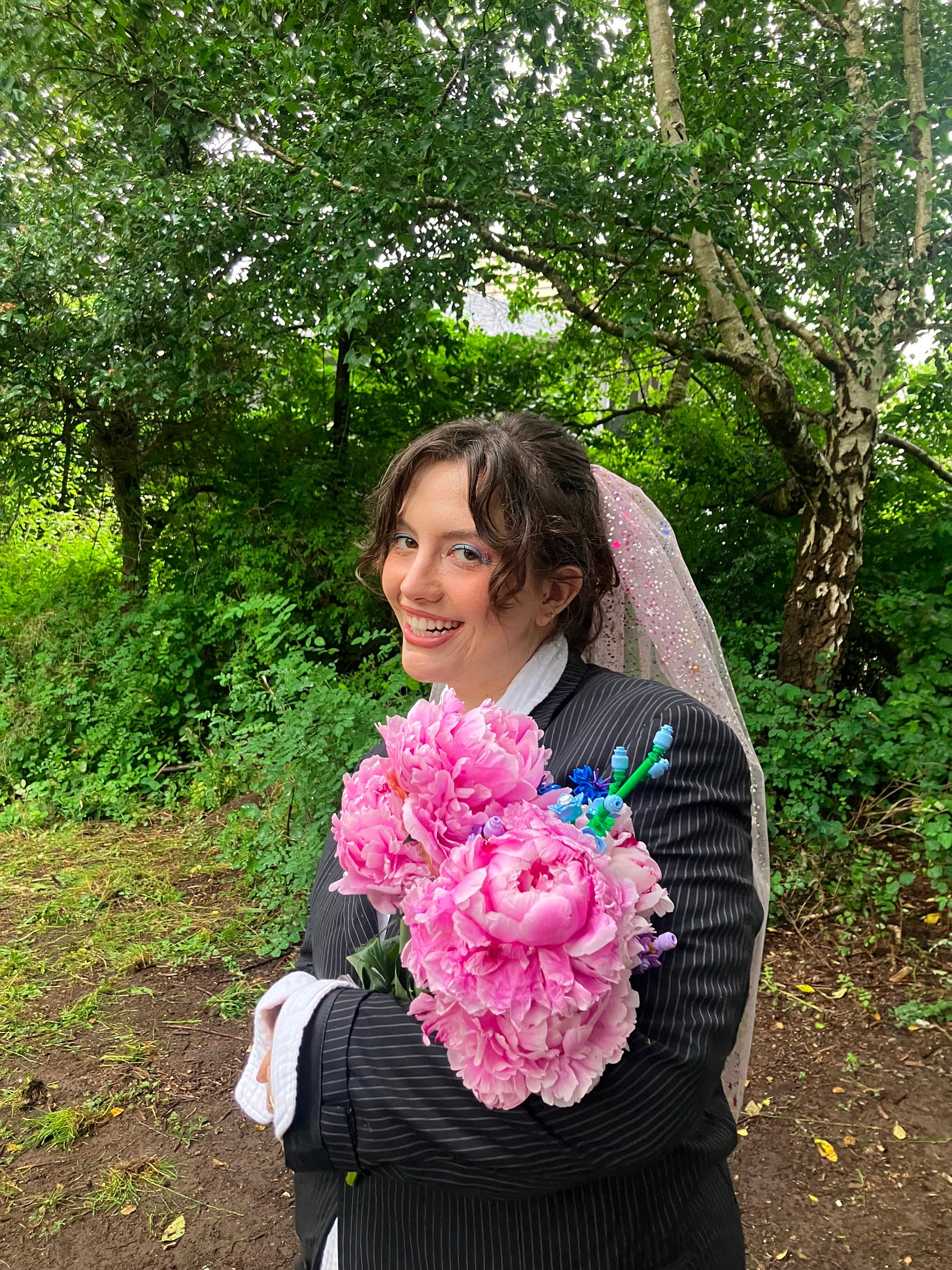 Photo of the author in a black blazer with a pink peony bouquet and pink veil on a green background with trees