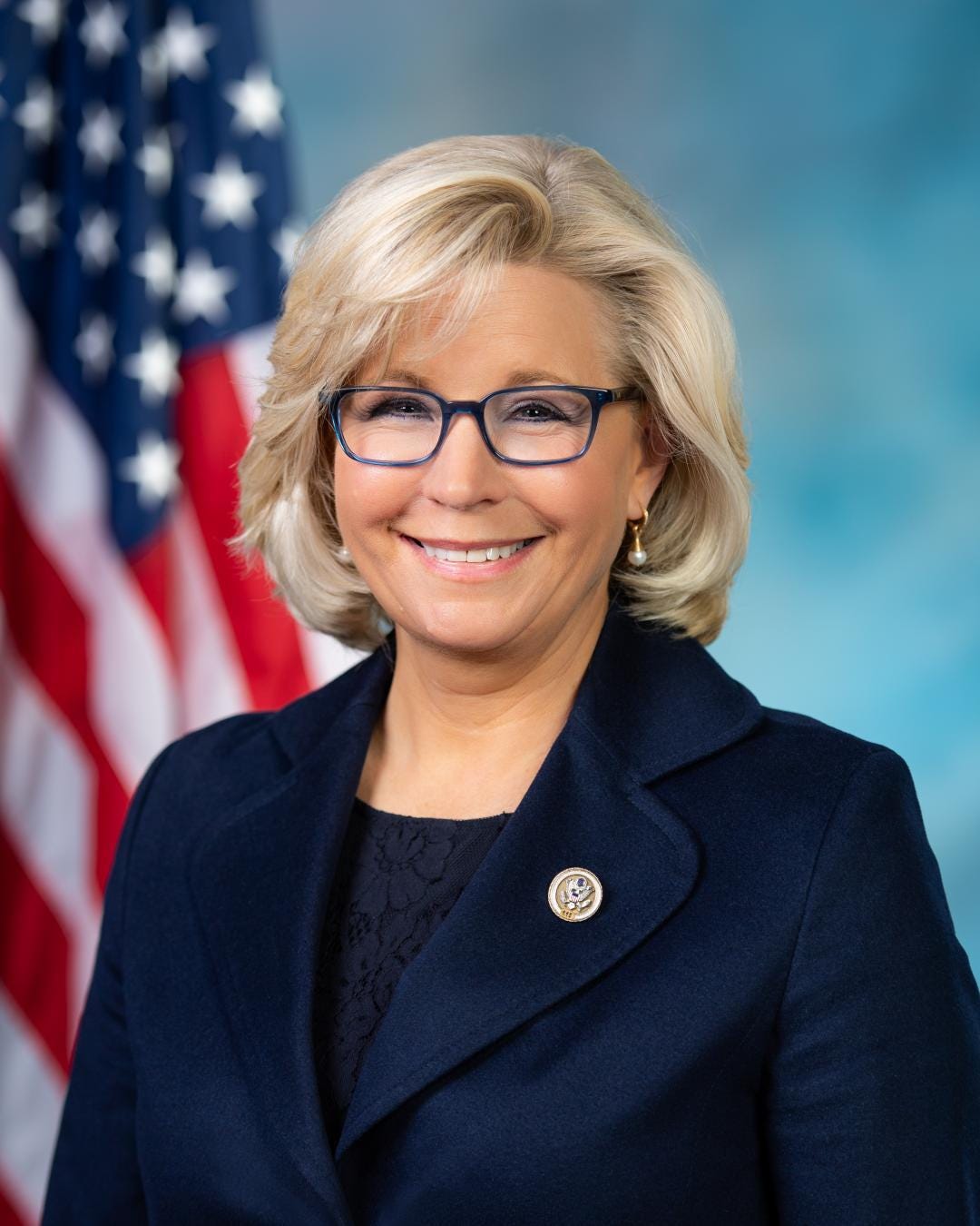 Liz Cheney, 2018 head and shoulder portrait, smiling woman in front of flag, blonde mid lengh hair, glasses, dark suit Liz Cheney, 2018 head and shoulder portrait, smiling woman in front of flag, blonde mid lengh hair, glasses, dark suit