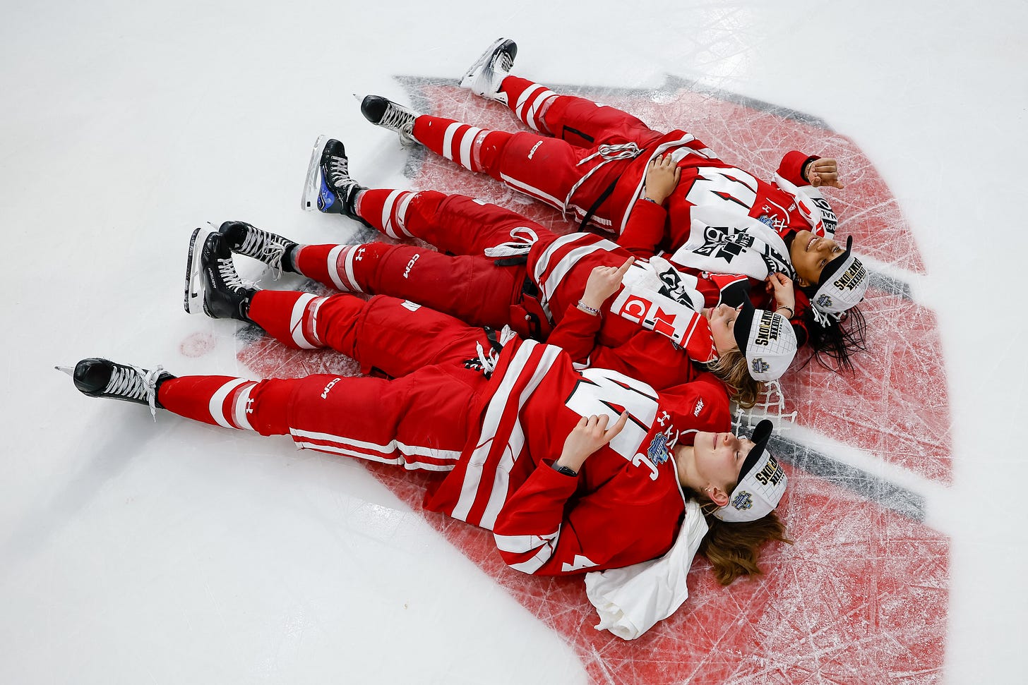 three Badger hockey players lay on the 'motion w' logo on the ice