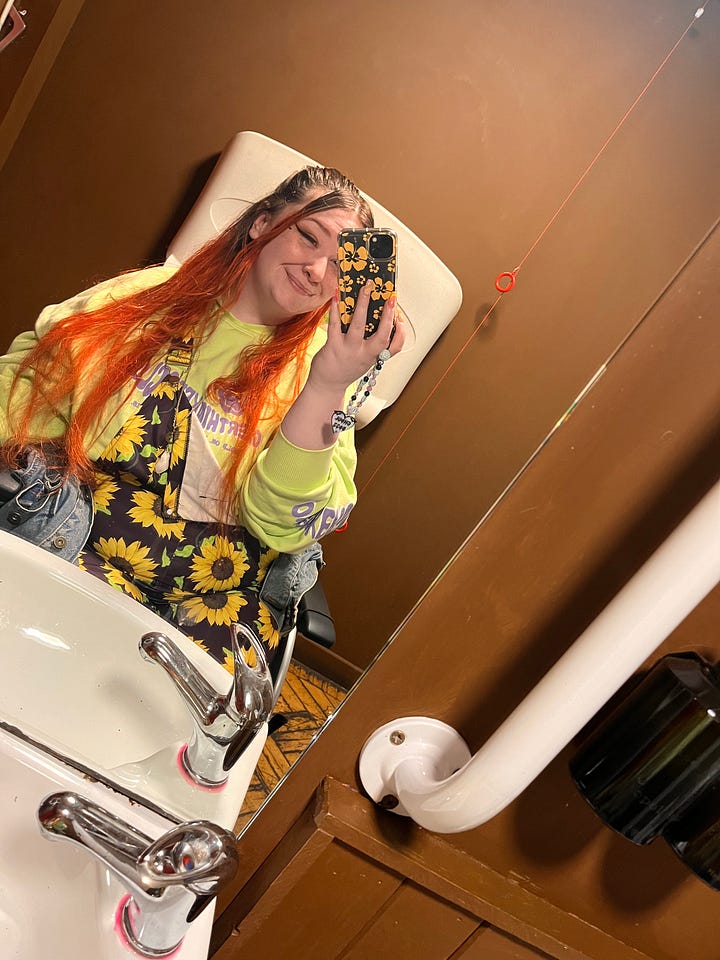 photo 1: A person seated in a wheelchair, smiling and holding up an iPad showing their career journey. They’re in a cosy room with yellow walls, autumn leaf decorations, and plants in the background.  photo 2: A mirror selfie in a hotel room. The person is wearing a bright orange jumpsuit with chain accessories, long orange hair, and giving a peace sign while seated in a wheelchair.  photo 3: A person sitting on a bed wearing a striped dress and a decorative tiara, smiling warmly. The room has colourful bedding, wall hangings, and soft string lights.  photo 4: A person in a wheelchair reaching through a fence to pet a Highland cow in a field. The scene is outdoors with fallen autumn leaves and trees with golden foliage.  photo 5: A mirror selfie taken in an accessible bathroom. The person is wearing sunflower-patterned clothing and a bright yellow top, smiling while seated in a wheelchair.