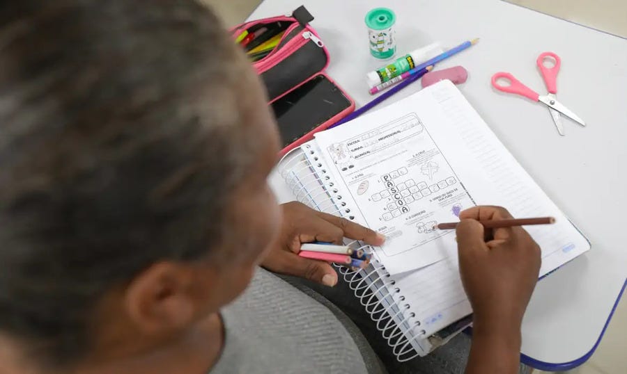 #descrevemospravocê  Pessoa sentada em uma mesa escolar realiza atividades em um caderno aberto. A fotografia é tirada de cima, em ângulo levemente diagonal, focando nas mãos da pessoa e no material sobre a mesa. Ela segura um lápis de cor marrom com a mão direita e desenha ou preenche uma atividade impressa no caderno, enquanto com a mão esquerda apoia a folha e segura dois lápis coloridos. A atividade parece envolver escrita e imagens relacionadas a palavras e letras, com figuras como bola e avião.  Sobre a mesa, ao lado do caderno, há uma tesoura com cabo rosa, uma borracha rosa, um tubo de cola com estampa de vaquinha, um lápis com ponteira azul e uma cola bastão. Também está visível um estojo preto com detalhes em rosa, parcialmente aberto, contendo outros materiais escolares. A pessoa usa camiseta cinza e tem cabelos presos. A imagem transmite concentração e dedicação a uma atividade de aprendizagem, possivelmente em um ambiente de alfabetização ou reforço escolar.