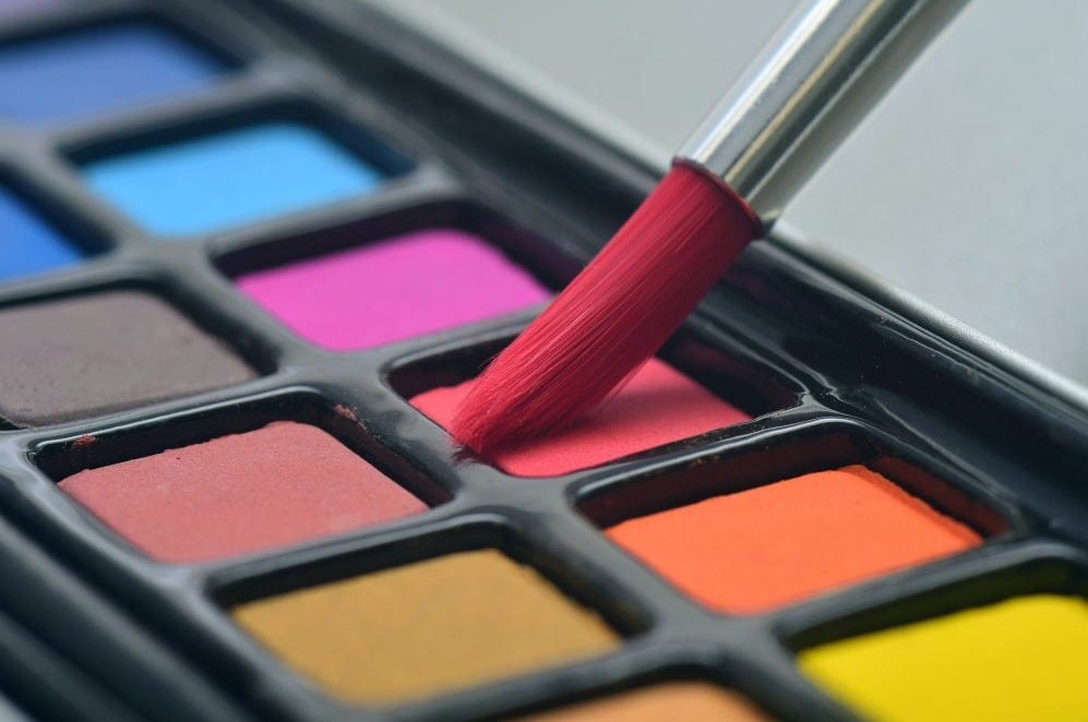 Red tipped paint brush hovering over the red paint square on a long black paint pallet with an array of colors. Red tipped paint brush hovering over the red paint square on a long black paint pallet with an array of colors.