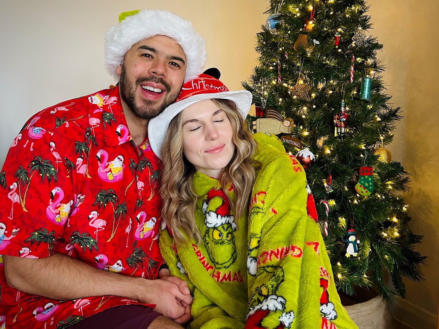 Jess. dressed in a Grinch blanket hoody and Christmas hat, sits next to Luke in front of a Christmas tree