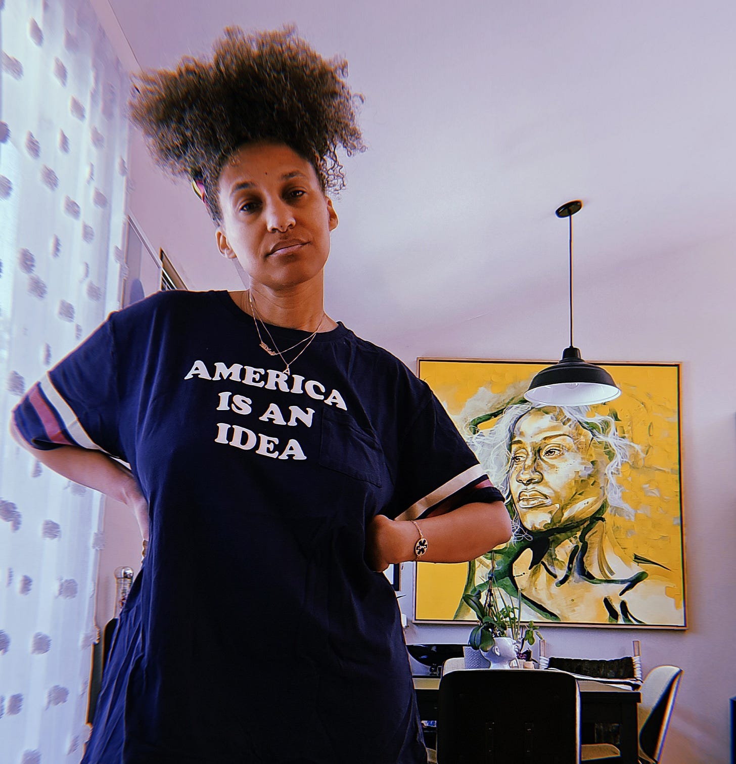 Dr. Cadet stands in her home with her hands on her hips wearing an oversized blue T-shirt that says AMERICA IS AN IDEA. With her head to the side and natural curls pinned on top of her head a yellow and green painting of her by Taylor Smalls is on the wall behind her.
