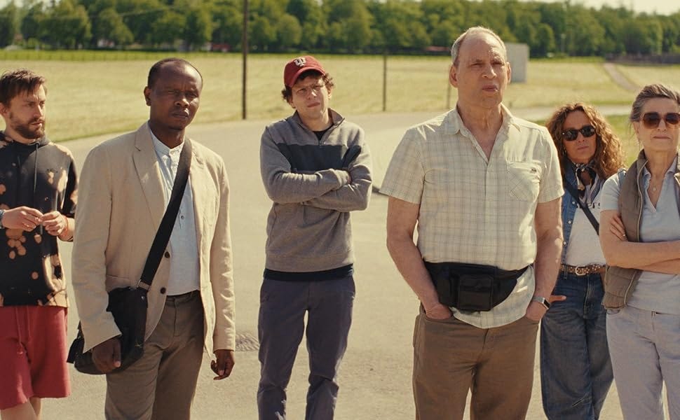 Still from the 2024 film A Real Pain. A group of six people stands outdoors in a rural setting on a sunny day, with an open field and trees in the background. The characters vary in expressions, from contemplative to neutral. At the center, a man wearing a light plaid shirt and a black fanny pack looks serious, while another man in a red baseball cap and gray sweatshirt stands with his arms crossed. The group includes individuals dressed casually, with distinct personalities conveyed through their clothing and body language, hinting at the dynamic relationships between them.