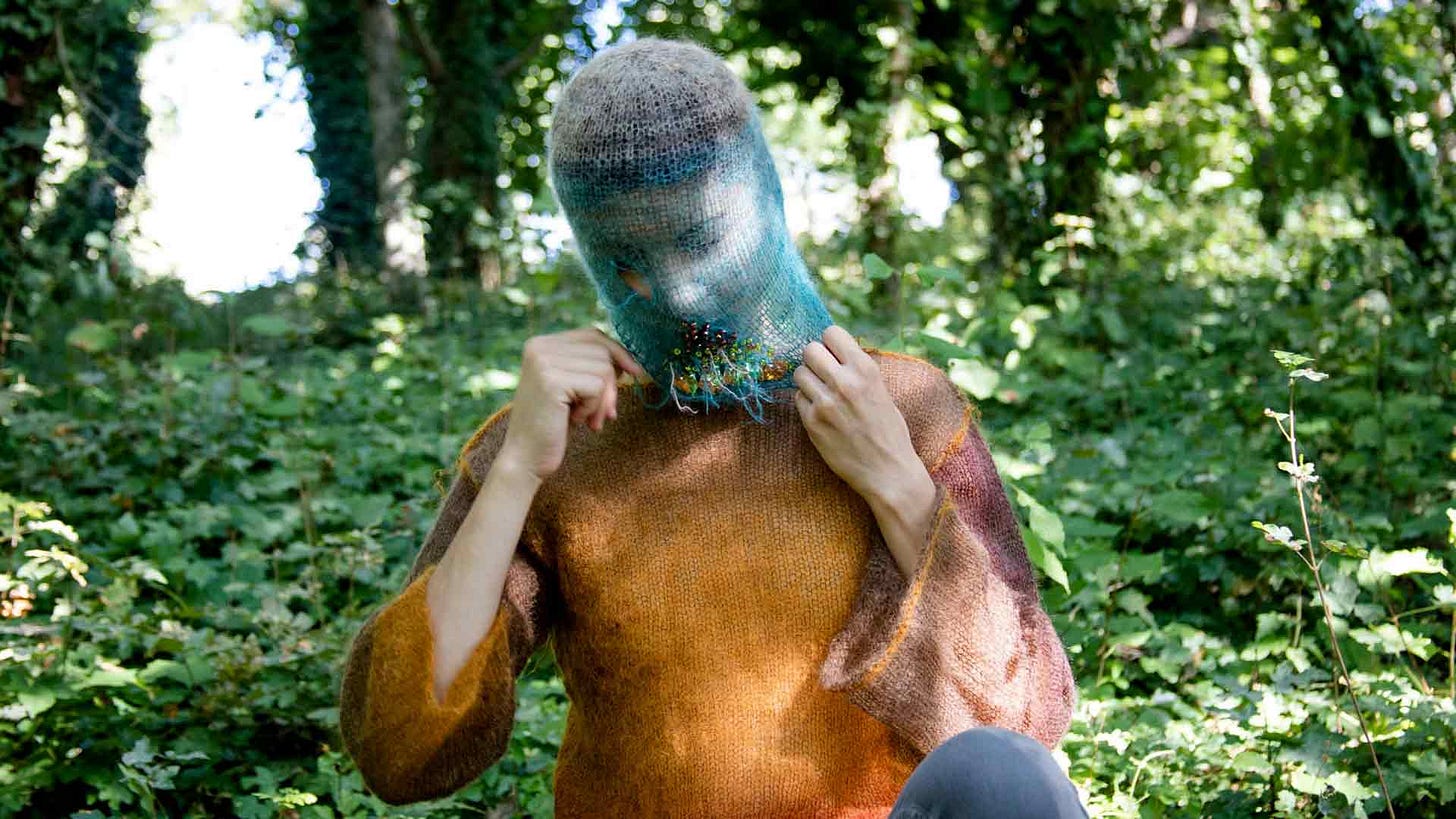 Imagem: Mulher segurando uma máscara azul feita com linha transparente e pedras no queixo, usando blusa laranja do mesmo tecido e uma floresta atras.