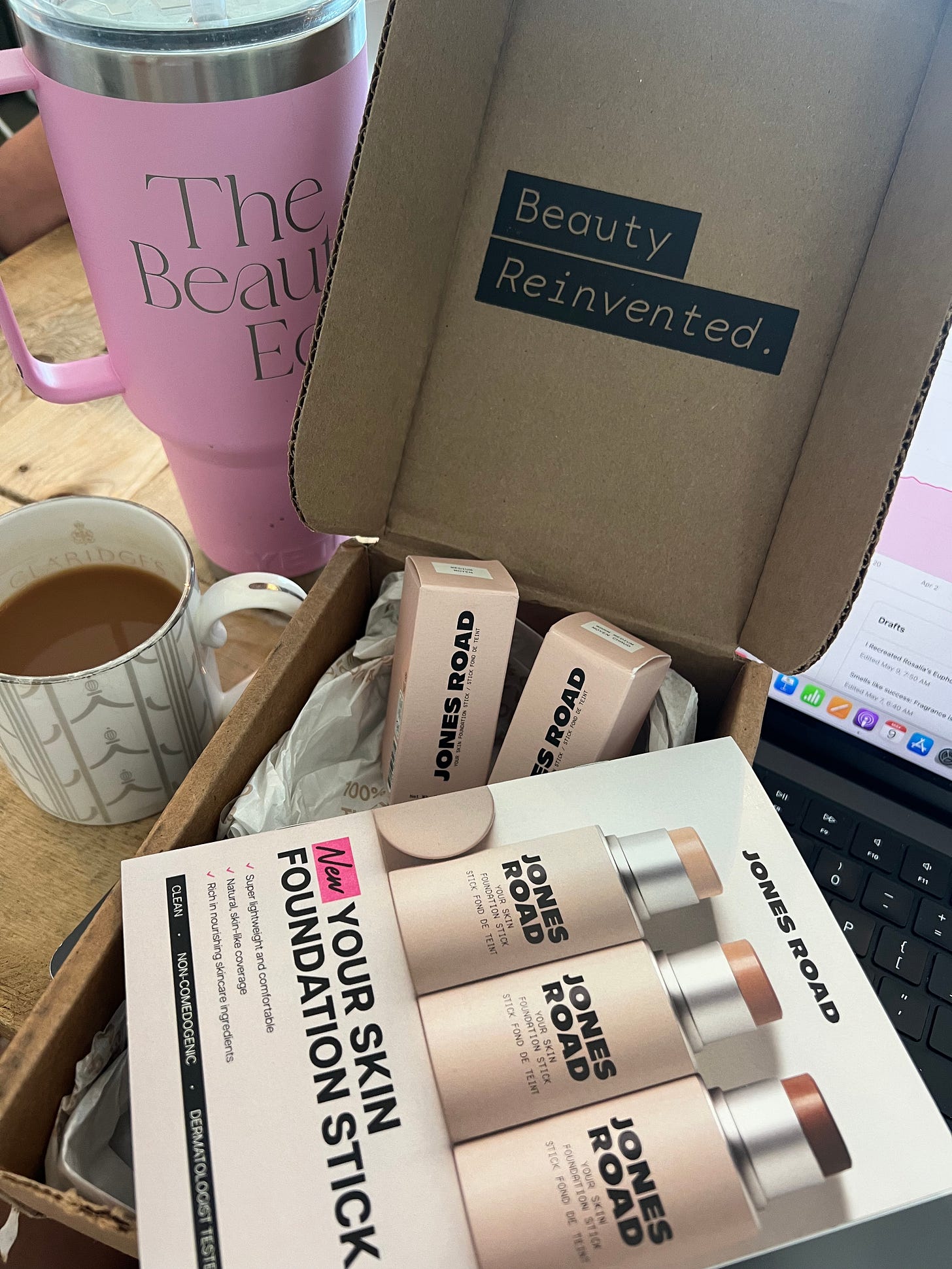 An open cardboard box labeled "Beauty Reinvented" contains two boxed Jones Road Foundation Sticks. In the background, a large pink tumbler with "The Beauty Ed®" logo sits next to a cup of tea and a laptop, creating a cozy "at-work" editorial setting.