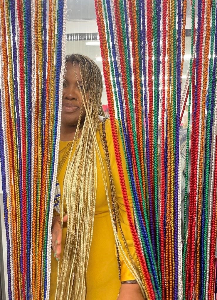A woman with long, braided hair is partially visible behind colorful strands of braided material. The strands are arranged vertically and feature a variety of bright colors, including red, green, blue, orange, and yellow. She is wearing a yellow top and has a hand reaching out, suggesting interaction with the colorful backdrop. The setting appears to be indoors, with soft lighting illuminating the scene.