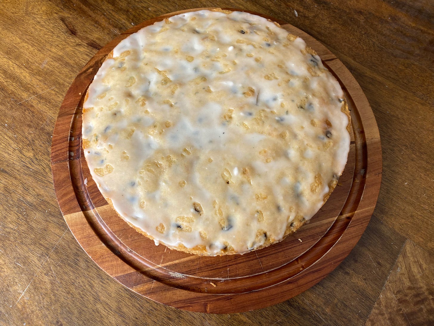 A round cake drizzled  with a thin lemon glaze and studded with specks of rosemary sits on a wooden cake board.