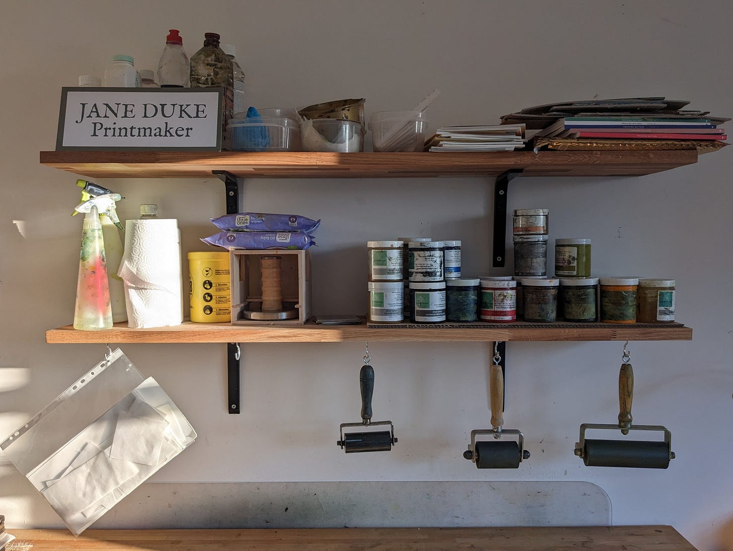 Two shelves bearing printmaking inks and materials. Ink rolers hang from hooks underneath. Two shelves bearing printmaking inks and materials. Ink rolers hang from hooks underneath.