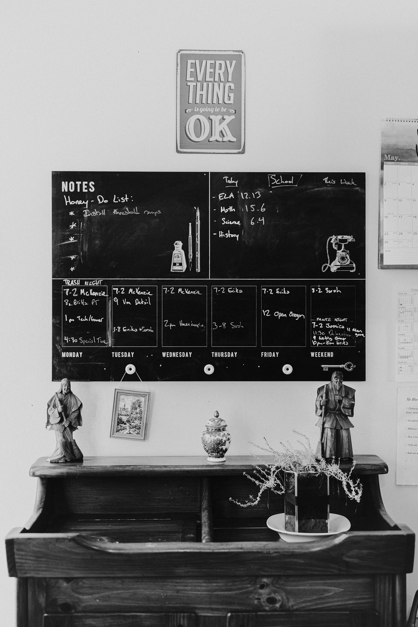 A sign stating “Everything is going to be OK” hangs above a messy chalkboard calendar with numerous details written in and a wooden dry sink with knickknacks on it. A sign stating “Everything is going to be OK” hangs above a messy chalkboard calendar with numerous details written in and a wooden dry sink with knickknacks on it.