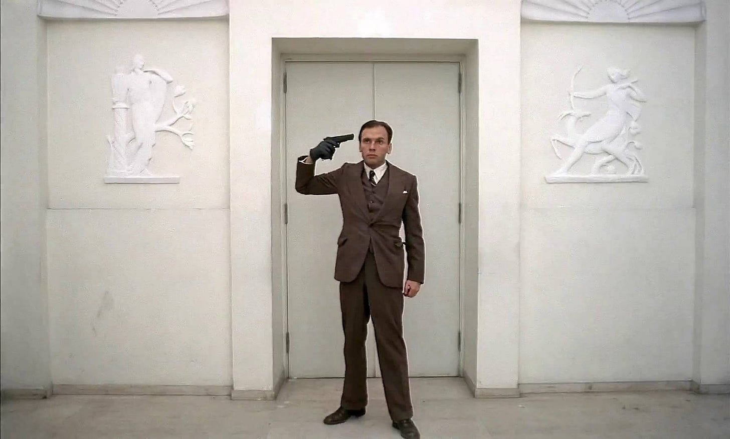 Jean-Louis Trintignant wears a suit and holds a pistol to his temple, a look of confusion on his face.