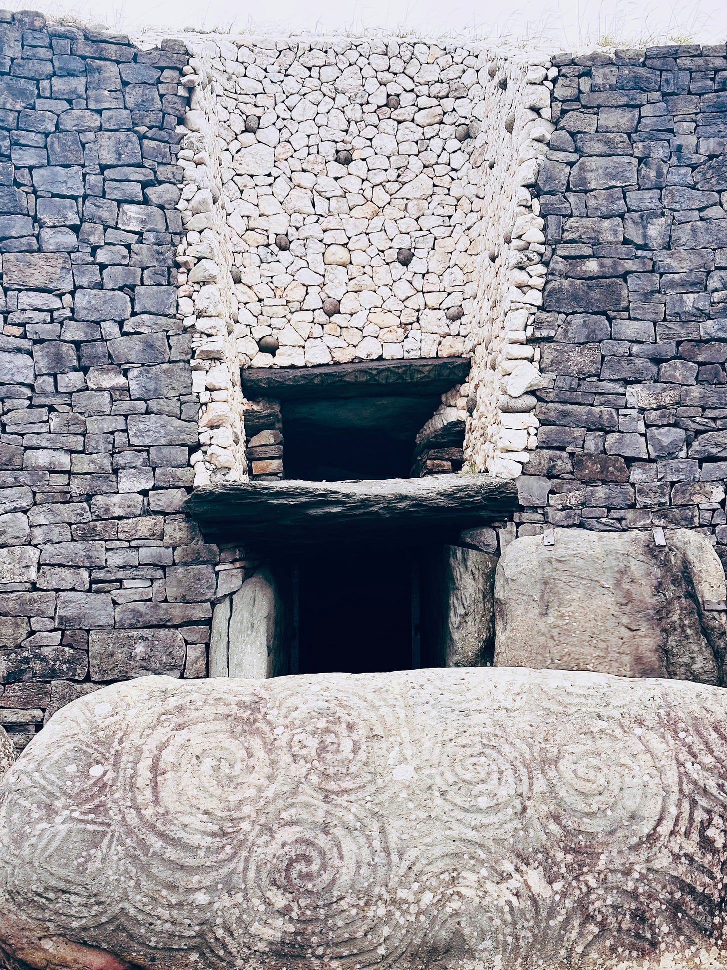 Photo of the front of Newgrange passage tomb. The facade is grey granite stones and white quartz stones stacked up on each other about 20 yards high. There is a door opening and a window opening above the door framed by flat stones. There is a large oblong stone in the foreground with spiral carvings on it. 